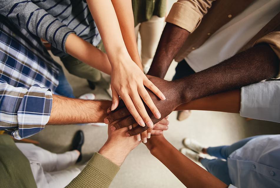 A top-down view of multiple people stacking their hands in the center of a team huddle, showing diverse skin tones and clothing in a collaborative setting.