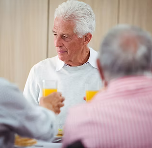 An elderly man with white hair sitting at a table, holding a glass of orange juice, with other people in the foreground and a softly lit indoor background.