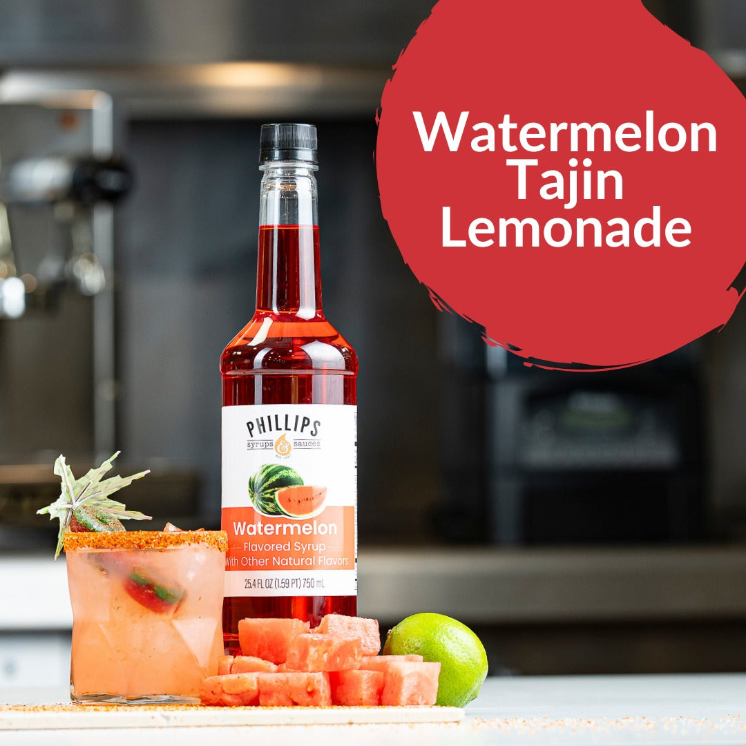 A bottle of Phillips Watermelon Tajin Lemonade syrup stands on a countertop with a prepared pink drink, watermelon cubes, and a lime in a blurred kitchen setting. A red graphic bubble reads 'Watermelon Tajin Lemonade.