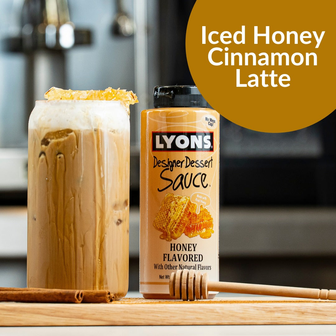 A bottle of Lyons Designer Dessert Sauce Honey Flavored shown next to an iced latte topped with syrup, on a wooden surface with a cinnamon stick and honey dipper.