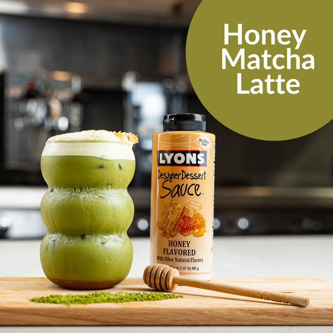 A honey-flavored bottle of Lyons Designer Dessert Sauce stands next to a green matcha latte in a textured glass on a wooden cutting board, with a honey dipper and matcha powder scattered in front. A green circle in the top right reads 'Honey Matcha Latte'.