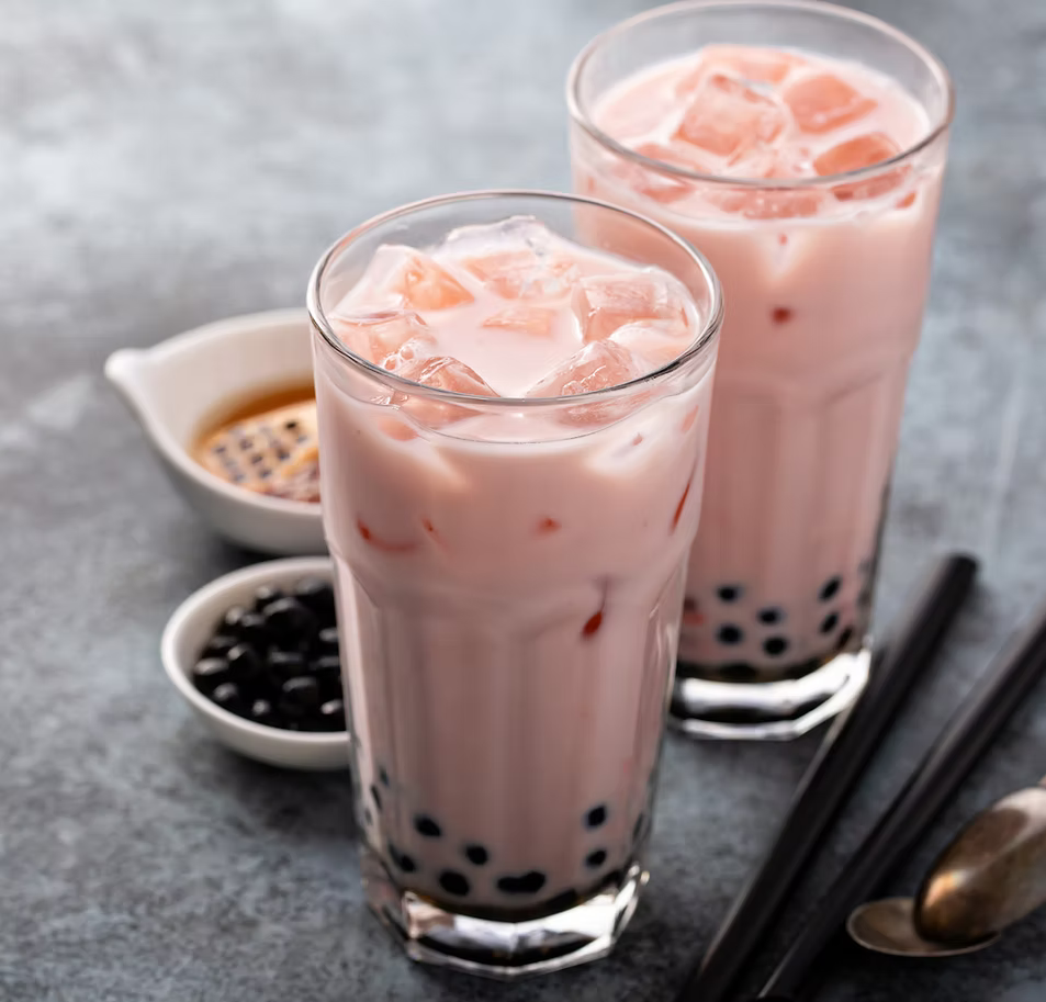 Two tall glasses of pink 珍珠奶茶 (boba tea) filled with ice cubes, set on a gray surface with small bowls of toppings, chopsticks, and a spoon.