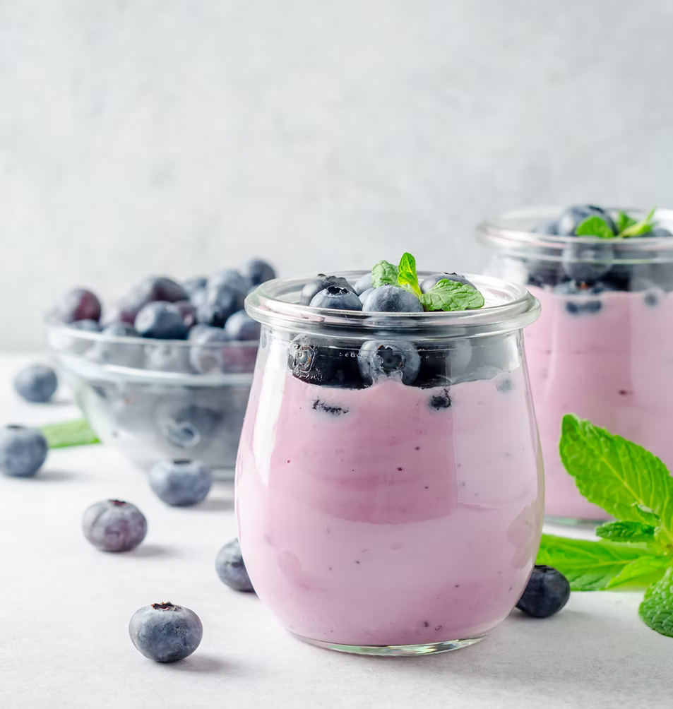 Two clear glass jars filled with pink berry yogurt, topped with fresh blueberries and mint leaves, set on a light surface with bowls of blueberries in the background.