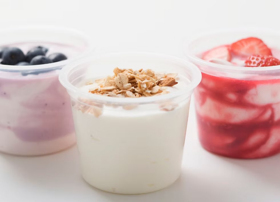 Three clear plastic cups of yogurt on a white surface, with the center cup filled with white yogurt topped with crumbled granola, flanked by purple berry yogurt with blueberries on the left and red strawberry yogurt with strawberries on the right.