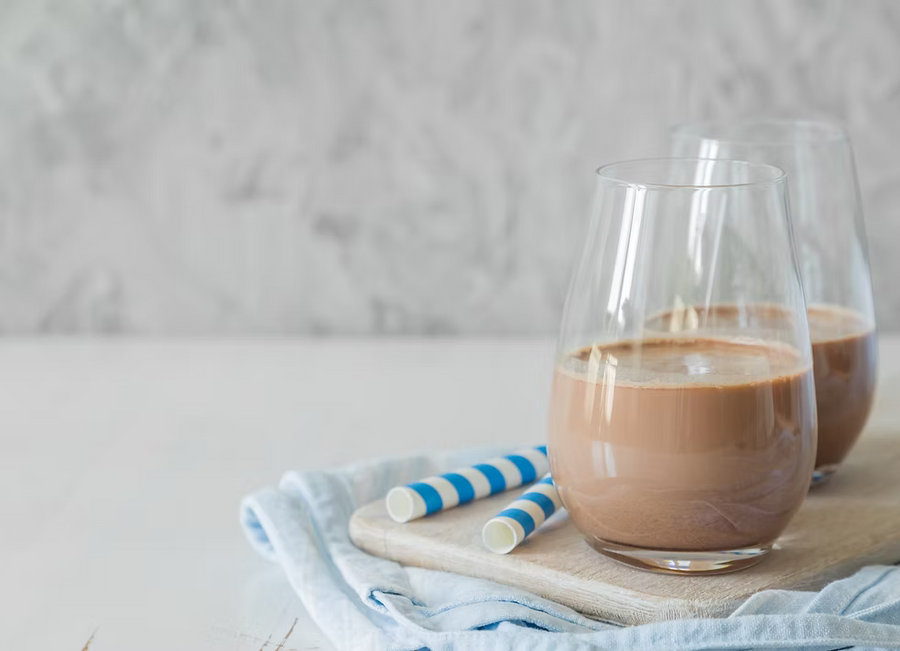 Two clear stemless wine glasses filled with a light brown iced coffee or milk-based drink, set on a wooden board with blue-and-white striped paper straws and a pale blue cloth against a soft light gray background.