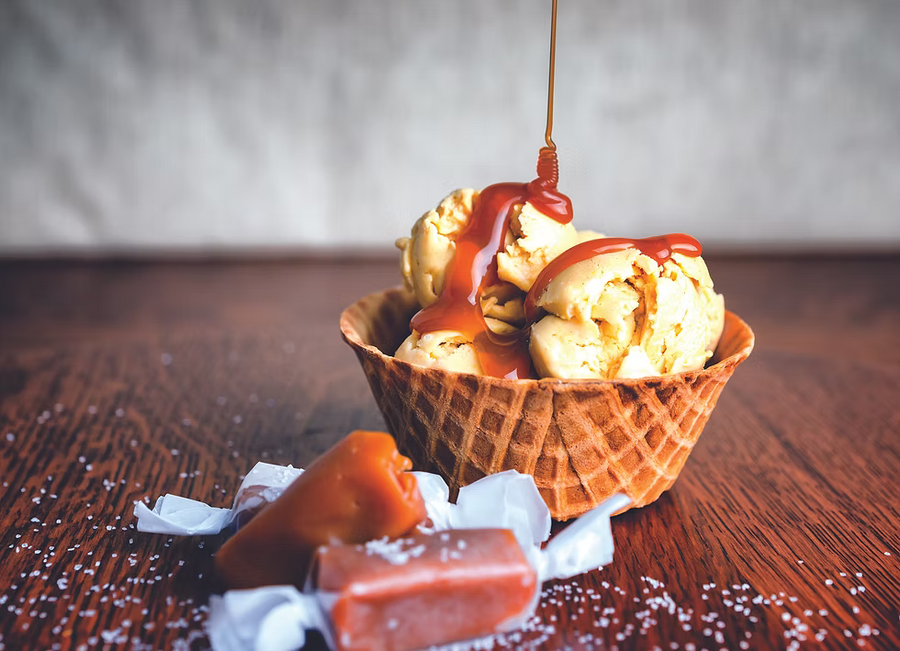 A waffle cone filled with vanilla ice cream and drizzled with caramel sauce, set on a dark wooden table with wrapped caramel candies in the foreground.