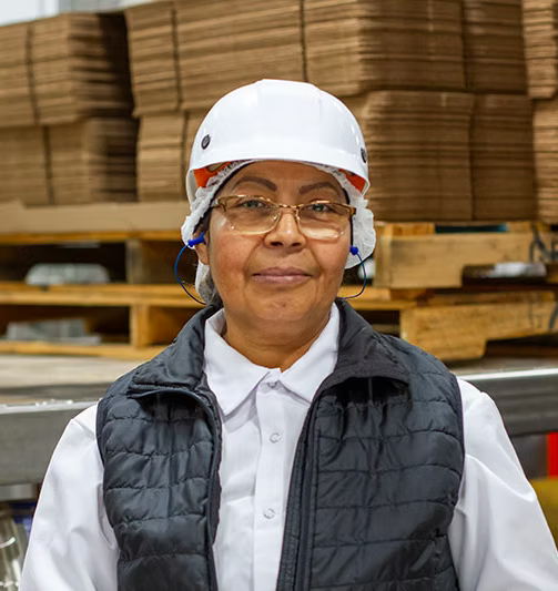 A woman wearing a white hard hat, safety glasses, a white shirt, and a black quilted vest stands in a warehouse or manufacturing setting with stacked cardboard boxes in the background.
