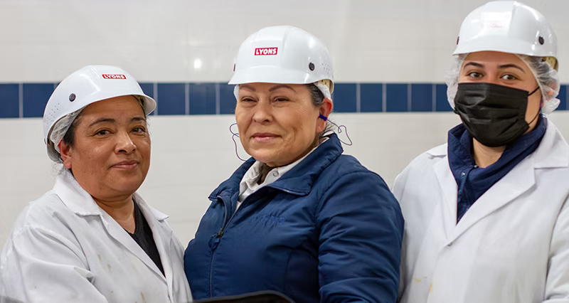 Three women wearing white hard hats with the LVFAB logo, white lab coats, and hairnets stand together in a tiled indoor setting. One woman on the right wears a black face mask.