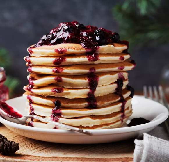 A tall stack of golden pancakes topped with a generous drizzle of dark berry sauce on a white plate, set on a wooden surface with a fork and spoon nearby.