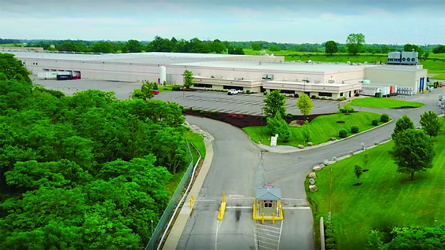 Aerial view of a large industrial warehouse building with a paved lot, access road, guard booth, and surrounding green trees and grass under a cloudy sky.
