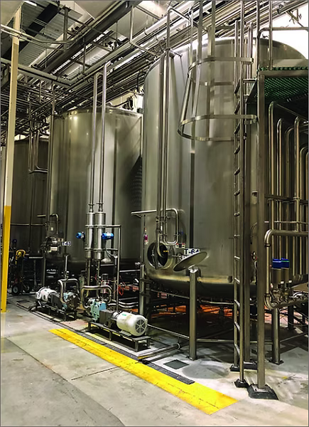 Large stainless steel fermentation tanks and piping systems in a brewery or industrial production facility, with concrete floors, yellow safety markings, and overhead utility pipes.