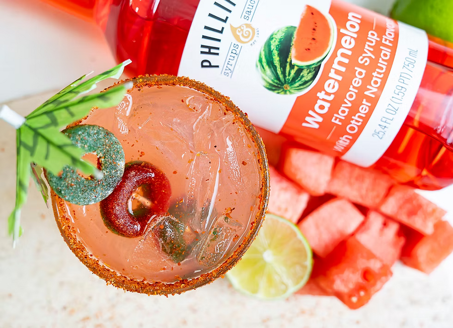 A glass of pink watermelon margarita with a lime wheel and jalapeño garnish, next to a bottle of Phillip's Syrup & Salsa Watermelon Flavored Syrup and watermelon cubes on a light surface.