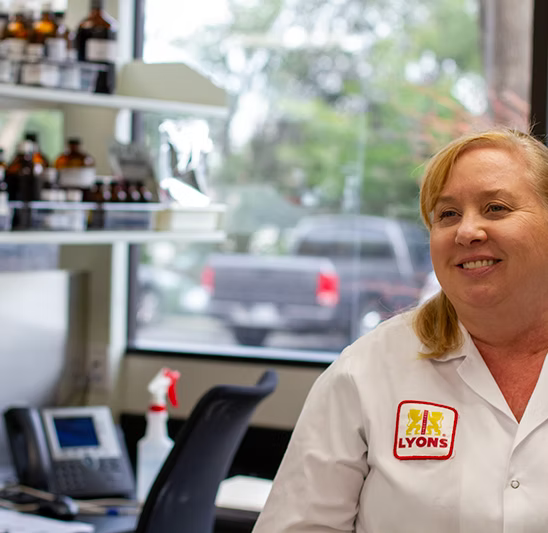 A smiling woman with blonde hair wearing a white Lyons brand lab coat stands in a laboratory setting with shelves of bottles and a window in the background.