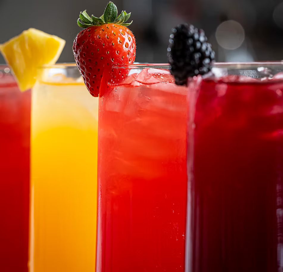 Three tall glasses of colorful iced cocktails in a row, garnished with a pineapple wedge, a strawberry, and a blackberry respectively, set against a blurred dark background.