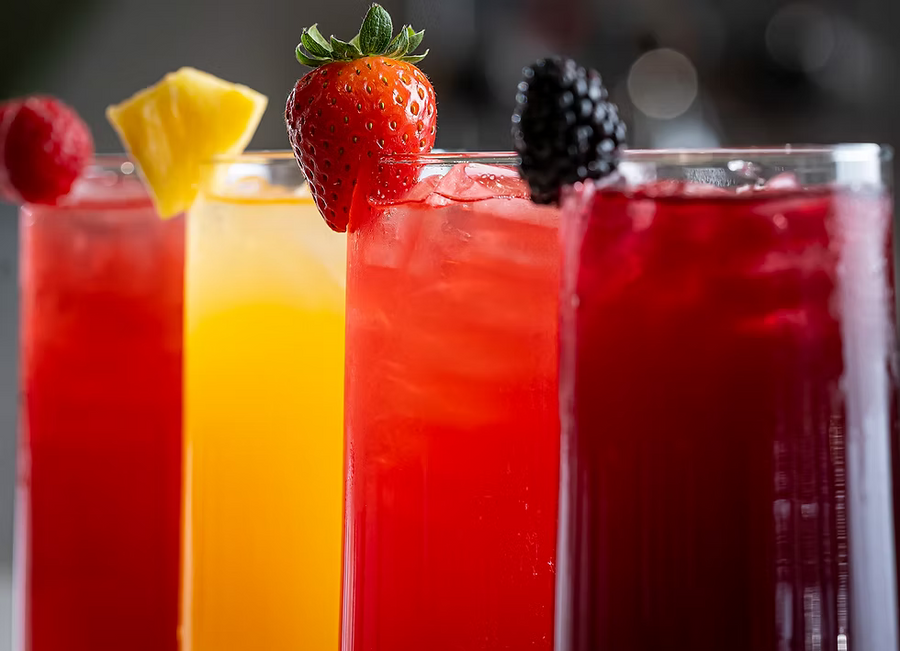 Four tall glasses of colorful iced cocktails lined up, garnished with fresh berries and citrus slices, set against a blurred dark background.