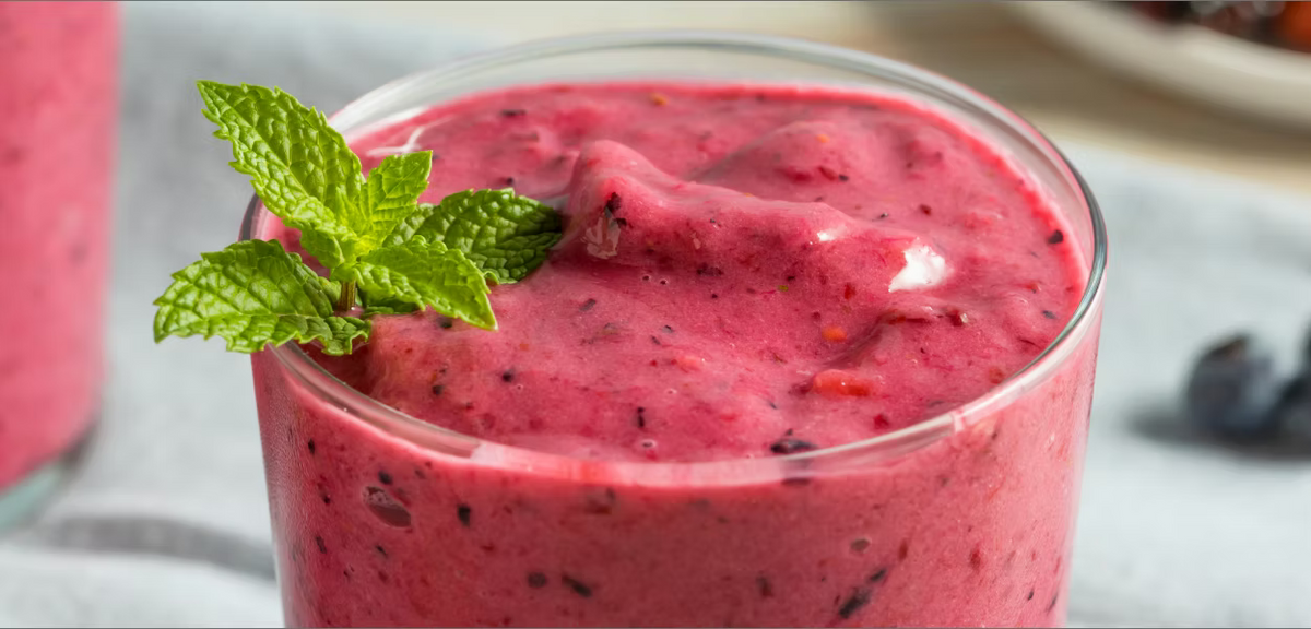 A close-up of a pink berry smoothie in a clear glass, garnished with a sprig of fresh green mint, with blurred blueberries on a light surface in the background.