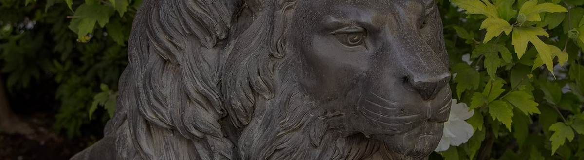 A close-up of a stone lion statue with a detailed mane and realistic facial features, set against a background of green foliage.