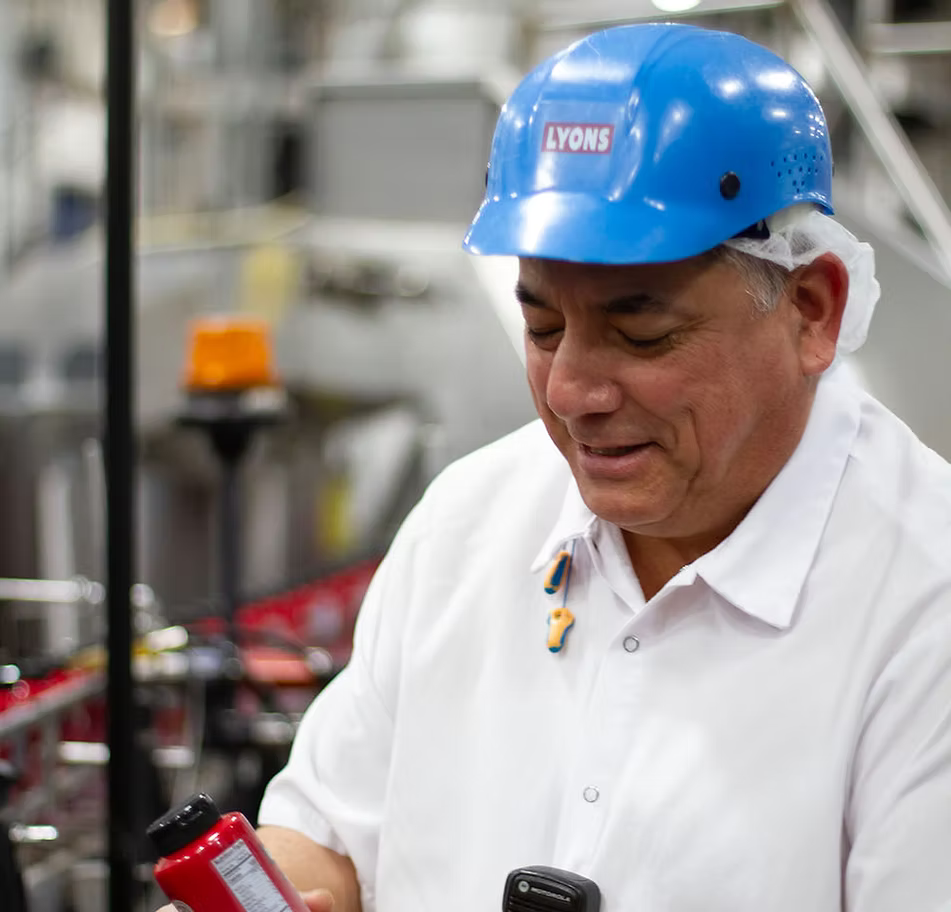 A man in a white shirt wearing a blue Lyons hard hat and hair cover is looking down at a red bottle in a clean, industrial setting.