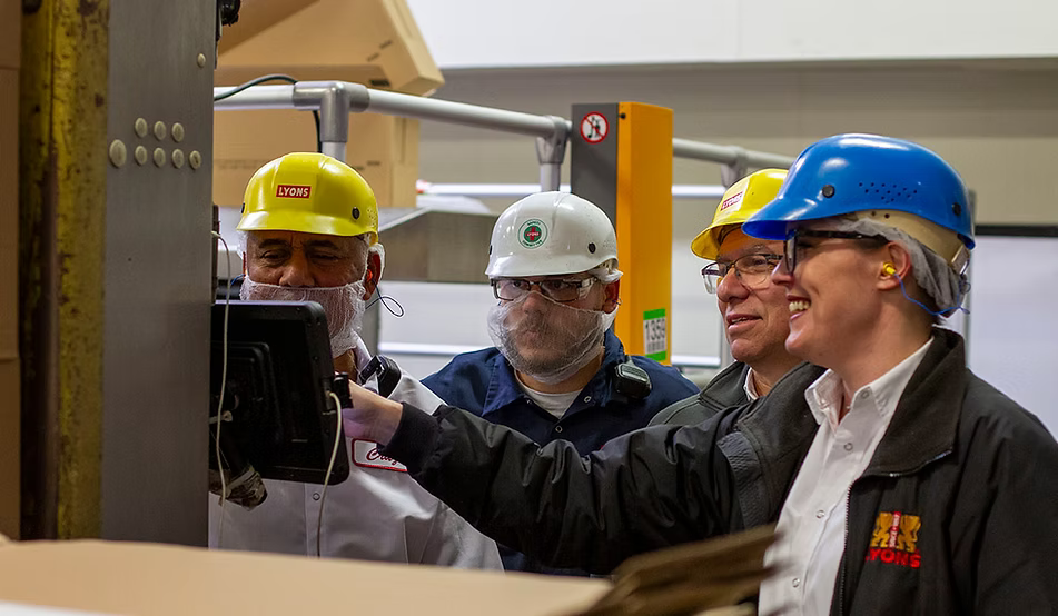 A group of workers wearing hard hats and face masks observe a screen in a factory setting. One person in a blue hard hat points toward the display.