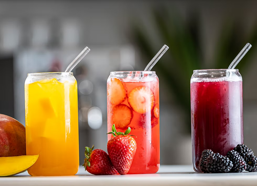 Three tall clear glasses filled with colorful iced drinks, arranged on a countertop with fruit slices and whole berries. The left glass contains bright yellow-orange juice, the center glass contains red fruit juice with strawberry slices, and the right glass contains deep red berry juice.