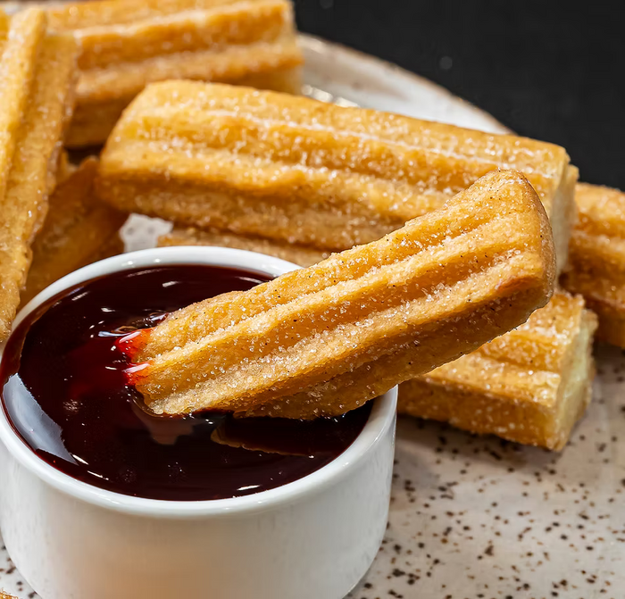 Crispy golden-brown churriegas dipped in dark chocolate sauce in a small white bowl, with more churriegas piled on a speckled plate.