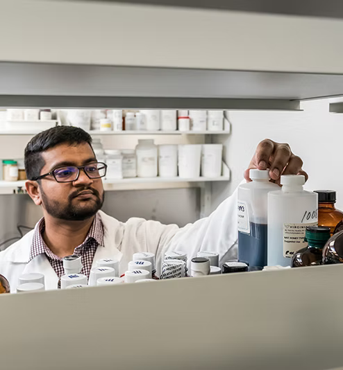 A man in a white lab coat and glasses looks thoughtfully while holding a white bottle in a laboratory setting filled with various scientific containers and supplies.