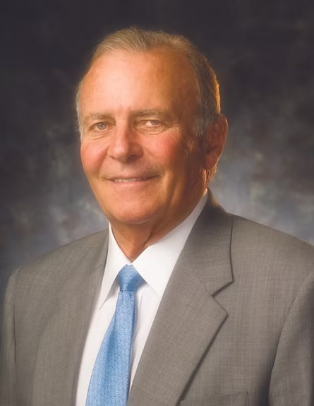 A professional portrait of an older man with gray hair, wearing a gray suit, white dress shirt, and light blue tie, set against a dark textured studio background.