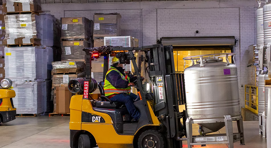 A yellow CAT forklift with a worker seated inside, positioned in a warehouse next to a large silver metal tank. Pallets and stacked goods are visible in the background.