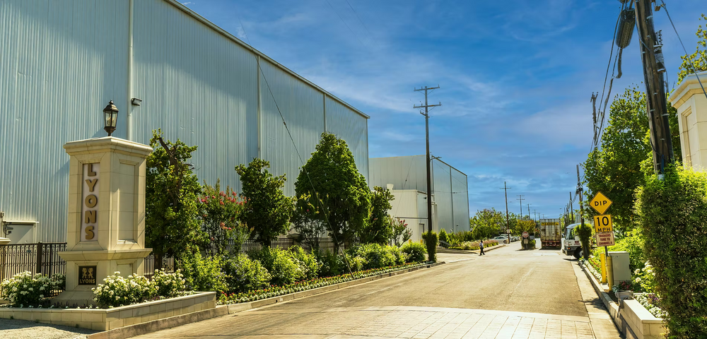 A wide outdoor street scene with a large light blue industrial warehouse building on the left, a paved road leading into the distance, green trees and shrubs along the property, utility poles, and a stone signpost with the name Lyons.