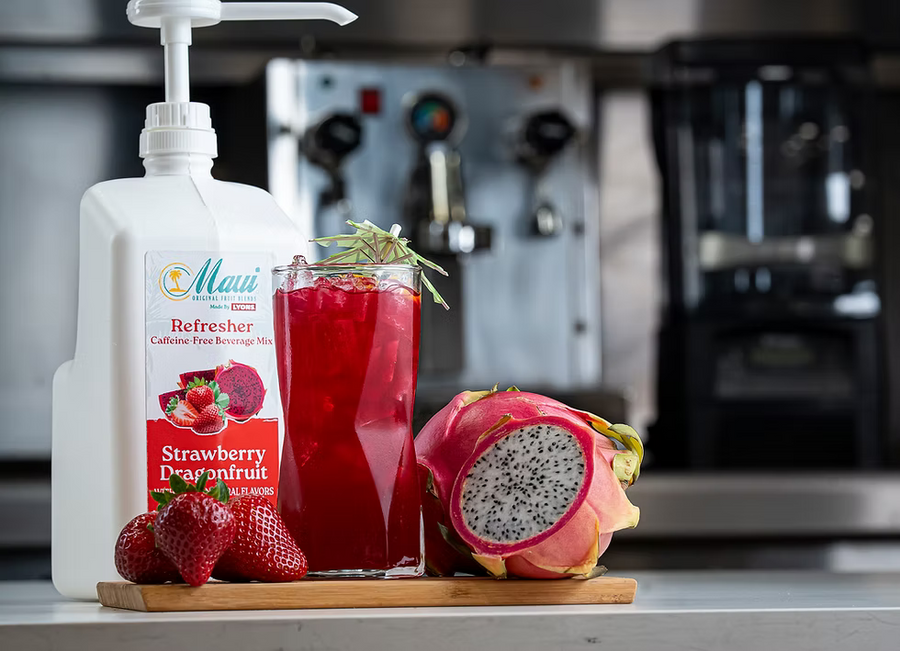 A bottle of Maui Refresher Strawberry Dragonfruit caffeine-free beverage mix stands on a counter beside a glass of bright red drink, fresh strawberries, a sliced dragonfruit, and a blurred kitchen background with a coffee machine.
