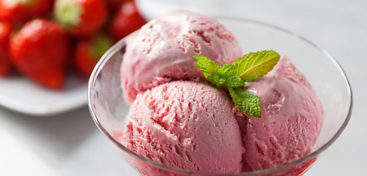 Three scoops of pink strawberry ice cream in a clear glass bowl, garnished with a mint leaf, with fresh strawberries blurred in the background.
