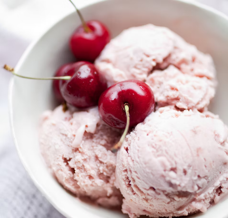 Scoops of light pink strawberry ice cream in a white bowl, garnished with three glossy red cherries on a softly blurred background.