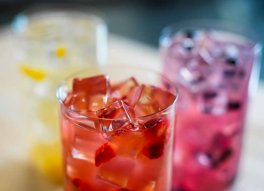 Three clear glasses filled with iced drinks and fruit pieces, photographed in a softly blurred real environment. The front glass contains a translucent red drink with red fruit pieces, while the rear glasses contain pink and pale yellow drinks.