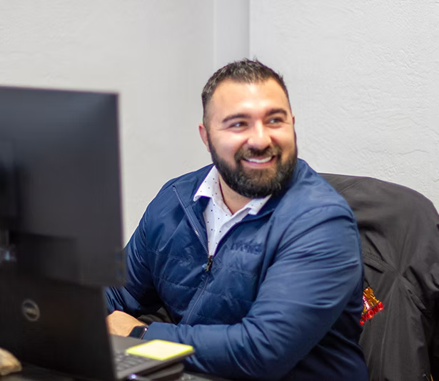 A smiling man with a beard wearing a blue zip-up jacket over a patterned collared shirt, sitting at a desk in front of a computer monitor.