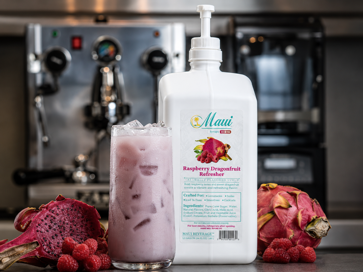 A large white bottle of Maui Raspberry Dragonfruit Refreshers syrup with a pump dispenser stands next to a tall glass filled with a pinkish-purple iced drink. Fresh dragonfruit halves and raspberries are arranged on both sides, with a blurred commercial kitchen background featuring espresso machines.