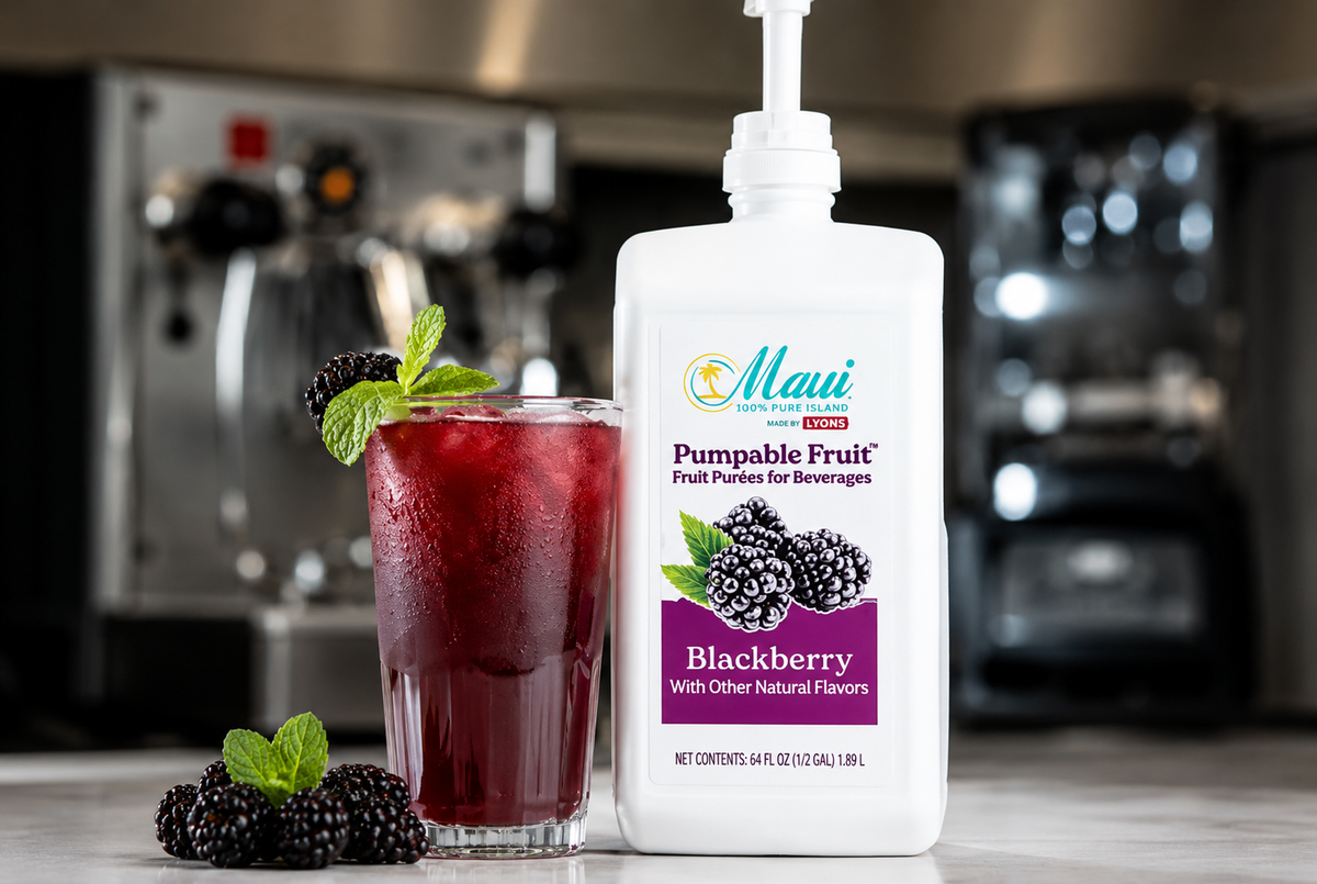 A bottle of Maui Pumpable Fruit Blackberry beverage mix stands on a countertop next to a tall glass filled with a dark red blackberry drink, garnished with mint, with a blurred kitchen background.