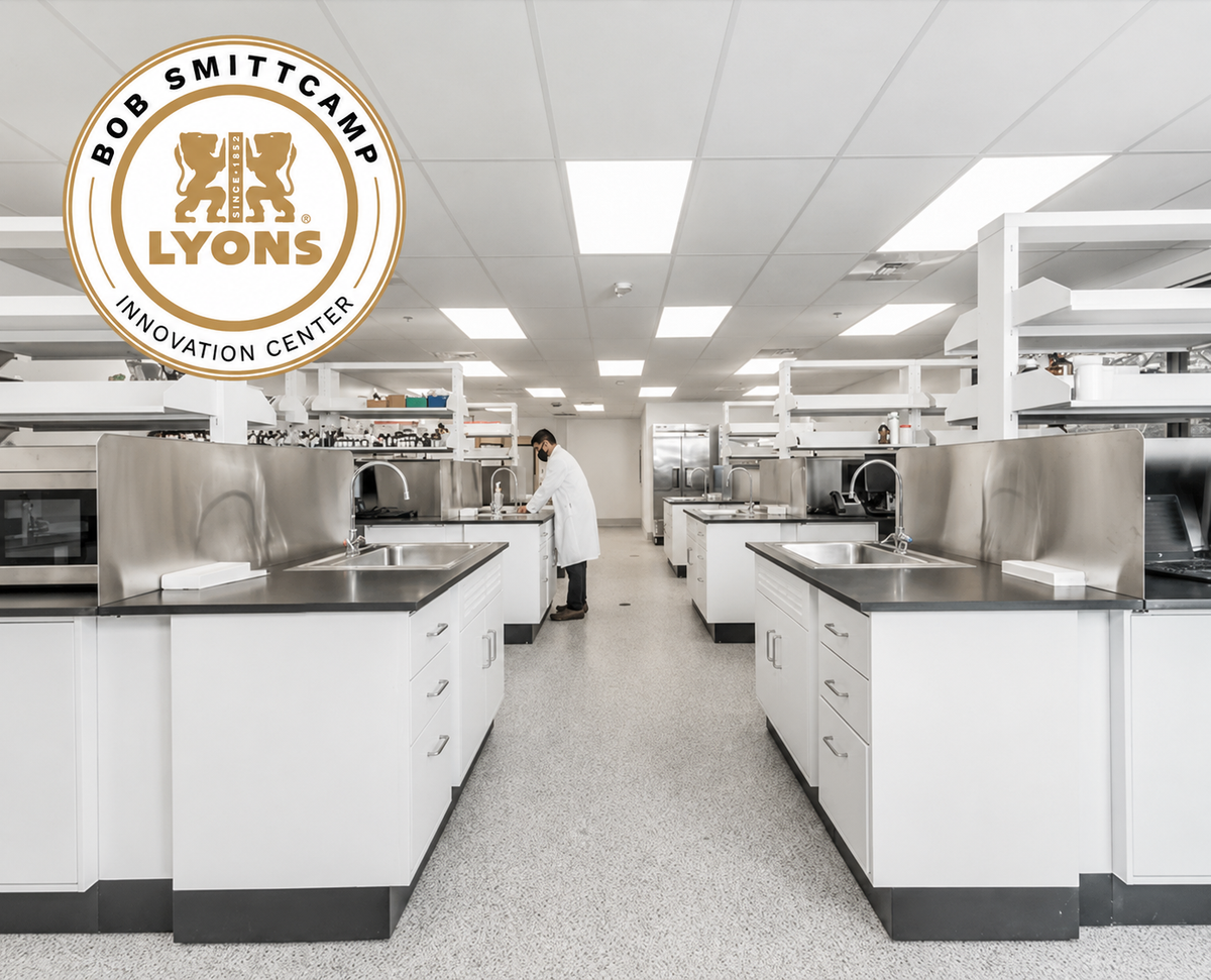 A wide interior view of a modern science lab with white cabinets, stainless steel work surfaces, bright ceiling lights, and a person in a white coat standing near the center aisle. A circular Bob Smittkamp Lyons Innovation Center logo is overlaid in the upper left.
